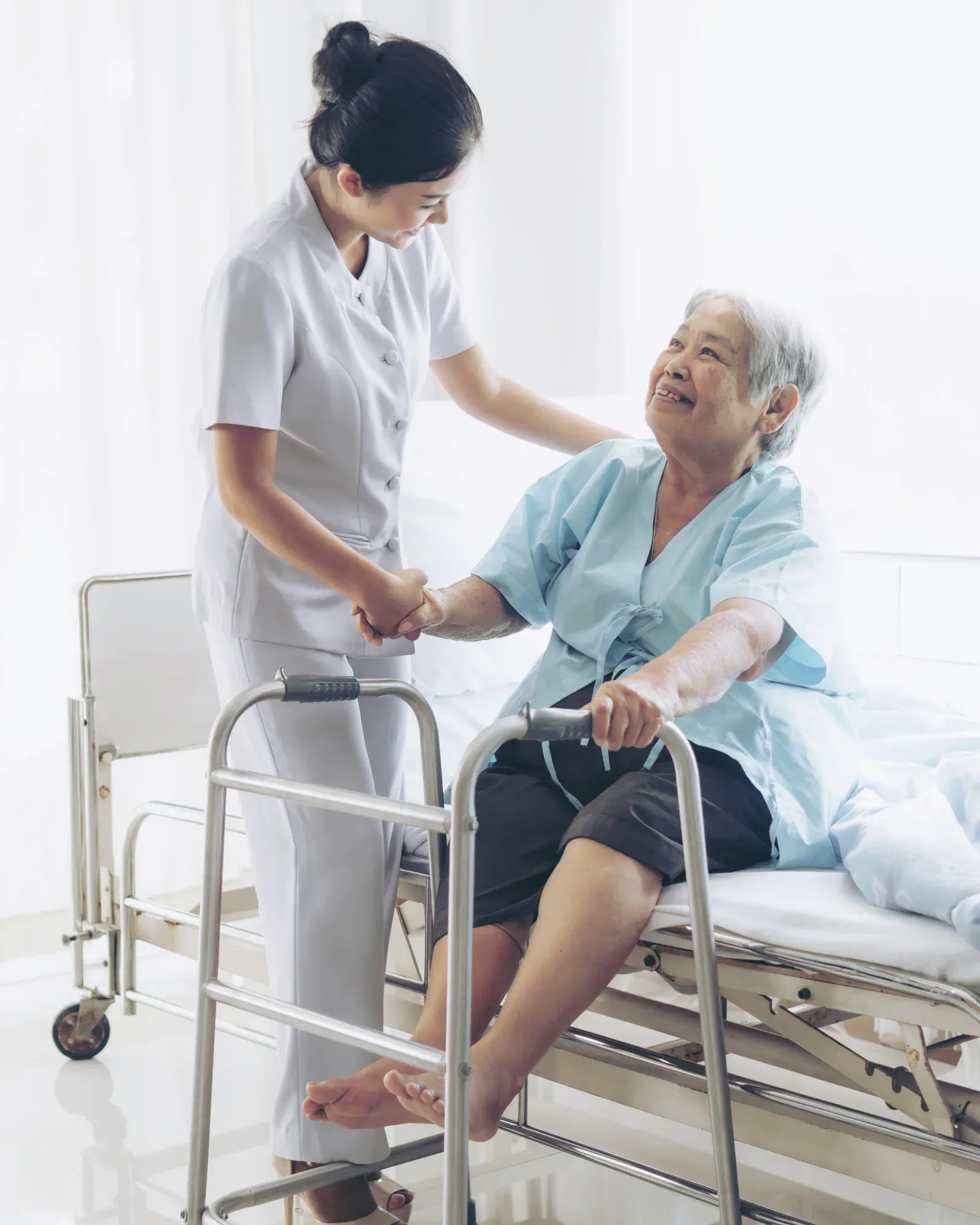 Una enfermera de uniforme blanco toma de la mano a una paciente mayor sentada en la cama de un hospital, junto a un andador; la paciente la mira sonriendo.